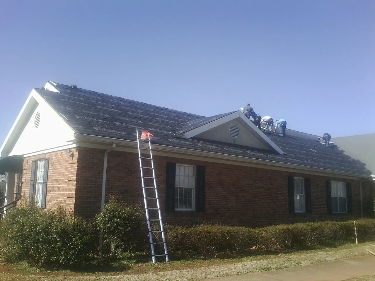 Crew tearing shingles off a brick ranch roof with a ladder set — Metal Roof Repair work by our crew in Alcove