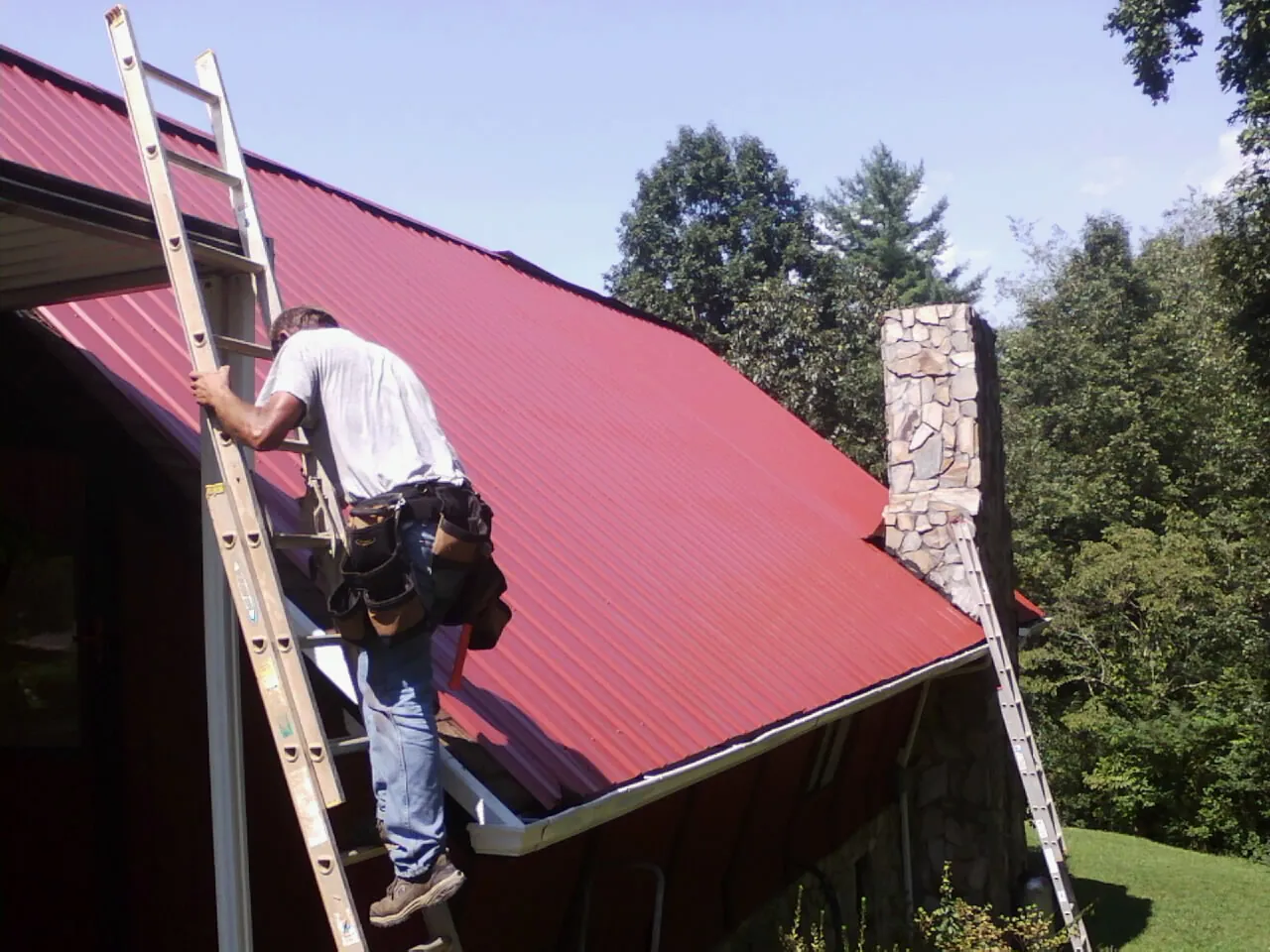 Roofer on a ladder beside a red metal roof and stone chimney — Soffit & Fascia Repair by Roofing Brothers in Alcove
